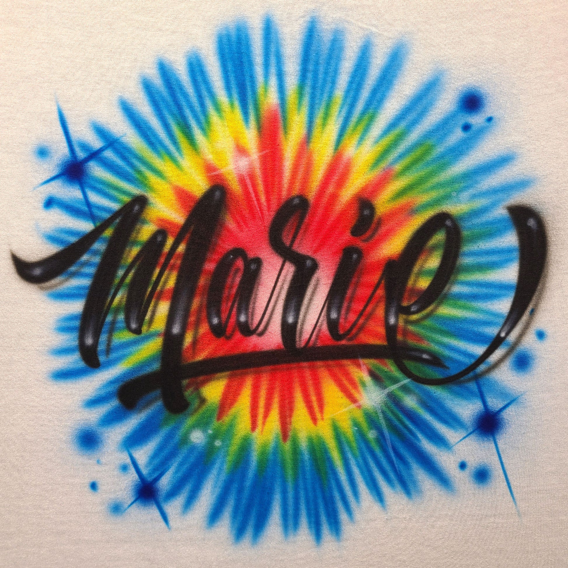 Custom airbrush shirt featuring bold black script lettering over a bright rainbow tie-dye burst in red, yellow, green, and blue.