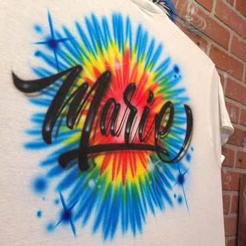 Custom airbrush shirt featuring bold black script lettering over a bright rainbow tie-dye burst in red, yellow, green, and blue.