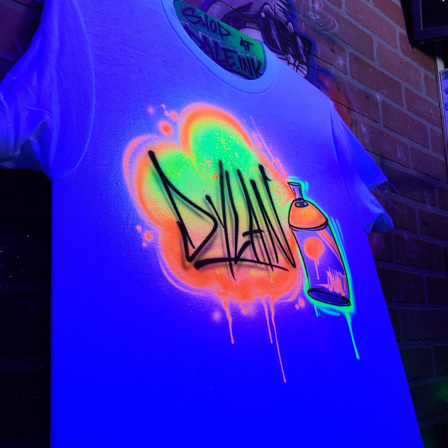 Custom glow-in-the-dark airbrush shirt with neon spray paint graffiti name "Dylan" and spray can artwork, illuminated under blacklight at AirbrushCustoms.