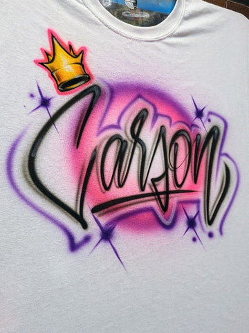 Custom airbrush shirt featuring the name "Carson" in bold black script lettering with a golden crown, pink and purple background, and star accents.