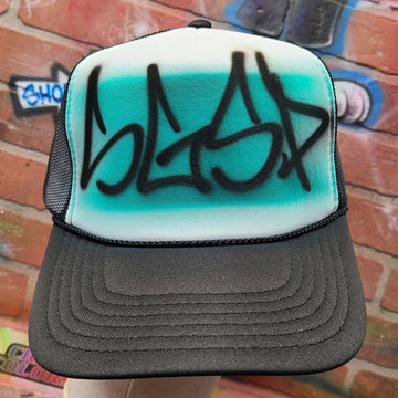 Custom graffiti airbrush hat with black handstyle lettering over a turquoise spray paint fade, on a black mesh trucker cap.
