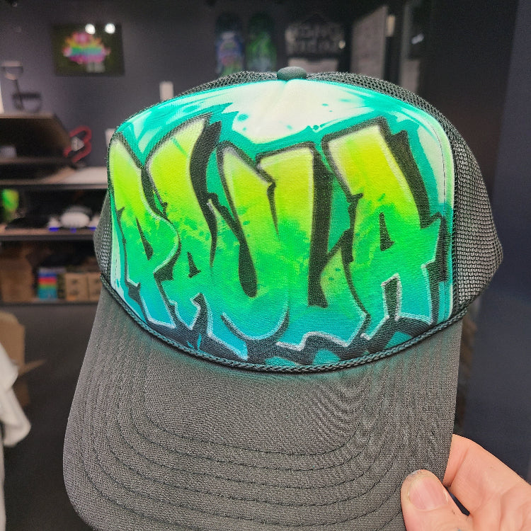 Custom airbrush hat featuring graffiti-style throwie bubble letters in green and teal fade with bold black outlines on a black mesh trucker cap.

