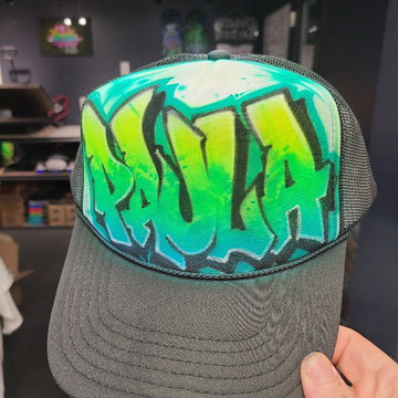 Custom airbrush hat featuring graffiti-style throwie bubble letters in green and teal fade with bold black outlines on a black mesh trucker cap.
