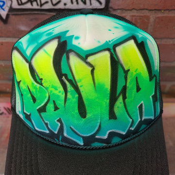 Custom airbrush hat featuring graffiti-style throwie bubble letters in green and teal fade with bold black outlines on a black mesh trucker cap.
