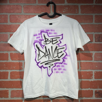 Airbrush shirt featuring the phrase “BE DANCE” in graffiti-style black lettering with a jagged purple outline and faded brick spray effect in the background.
