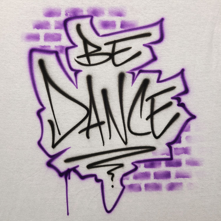 Airbrush shirt featuring the phrase “BE DANCE” in graffiti-style black lettering with a jagged purple outline and faded brick spray effect in the background.
