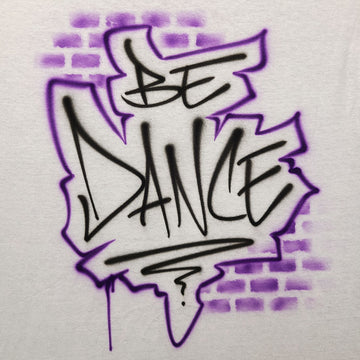 Airbrush shirt featuring the phrase “BE DANCE” in graffiti-style black lettering with a jagged purple outline and faded brick spray effect in the background.
