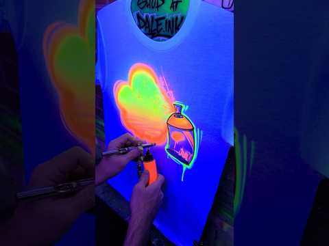 Video of a custom graffiti shirt being painted by an airbrush artist, showing the name “Dylan” in black spray-style lettering over a neon orange and green background, with a spray can graphic added in real time.