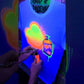 Video of a custom graffiti shirt being painted by an airbrush artist, showing the name “Dylan” in black spray-style lettering over a neon orange and green background, with a spray can graphic added in real time.