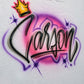 Custom airbrush shirt featuring the name "Carson" in bold black script lettering with a golden crown, pink and purple background, and star accents.
